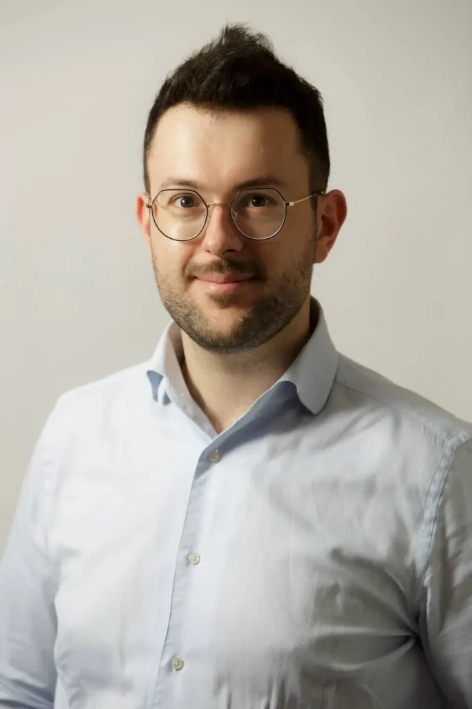 A man with short dark hair and glasses wears a light blue button-up shirt. He is facing the camera and smiling slightly, set against a plain light background.