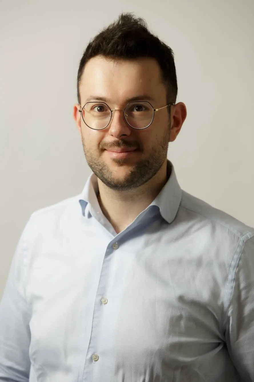 Un uomo con i capelli corti e scuri e la barba si erge sicuro su uno sfondo chiaro e semplice, indossando occhiali e una camicia azzurra abbottonata. Con un leggero sorriso, guarda verso la telecamera, incarnando la professionalità di qualcuno proveniente da una stimata azienda carni.