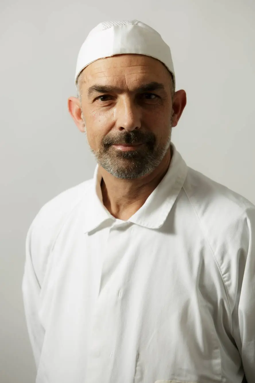 Un uomo che indossa un berretto bianco e un camice da cuoco, a nome di una rinomata azienda carni, si erge con sicurezza su uno sfondo semplice.