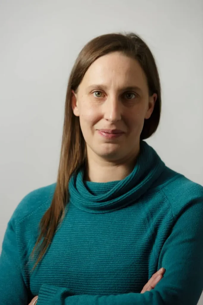 A woman with long brown hair, wearing a teal sweater, stands against a plain background. She has a neutral expression and her arms are crossed.