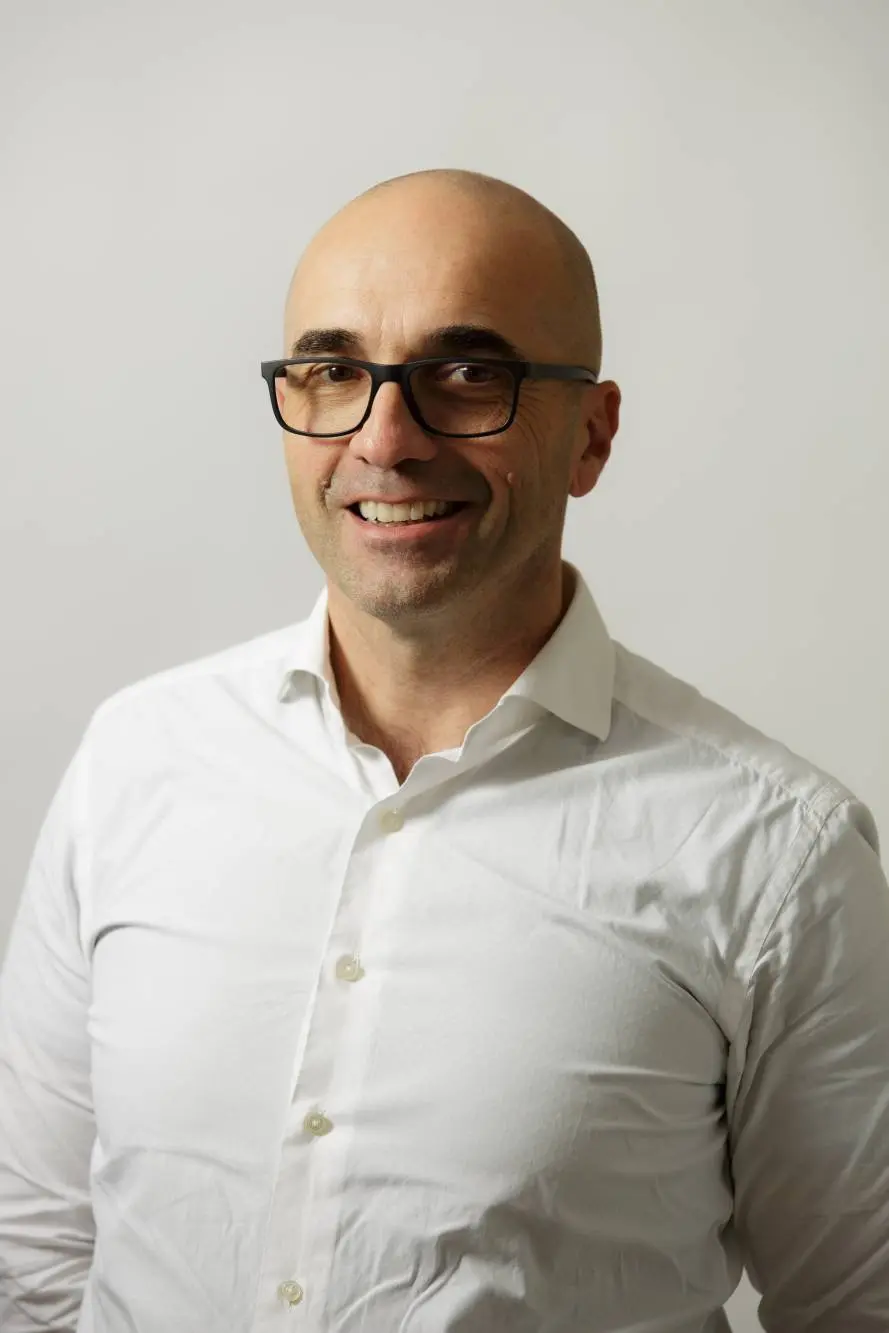 Una persona sorridente con gli occhiali e la testa rasata indossa una camicia bianca abbottonata, che trasuda la professionalità raffinata che si addice a un membro stimato dell'azienda carni. Sono in piedi su uno sfondo semplice e chiaro.