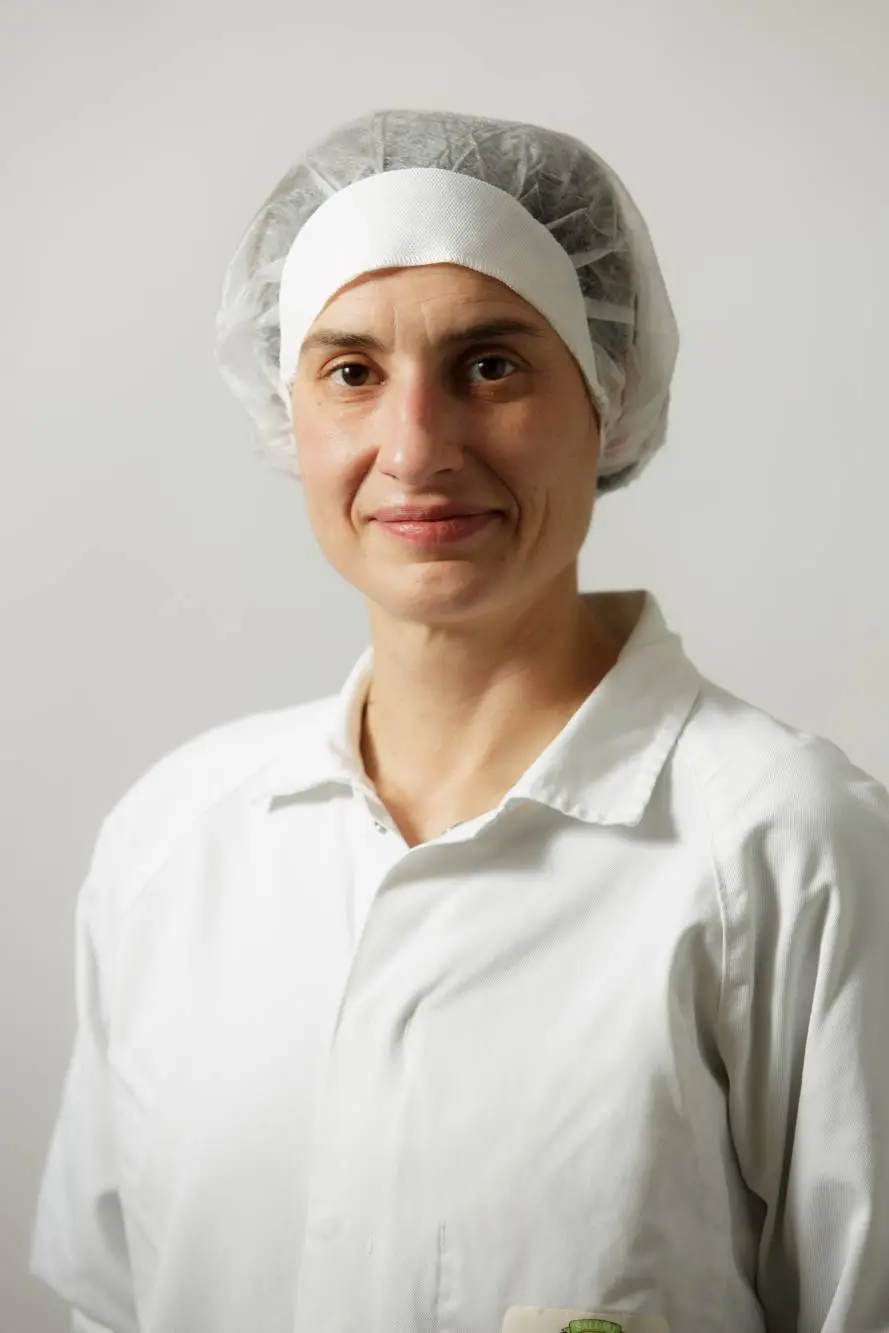 La persona che rappresenta l'Azienda Carni indossa un camice bianco e una retina per capelli e sorride leggermente su uno sfondo semplice.