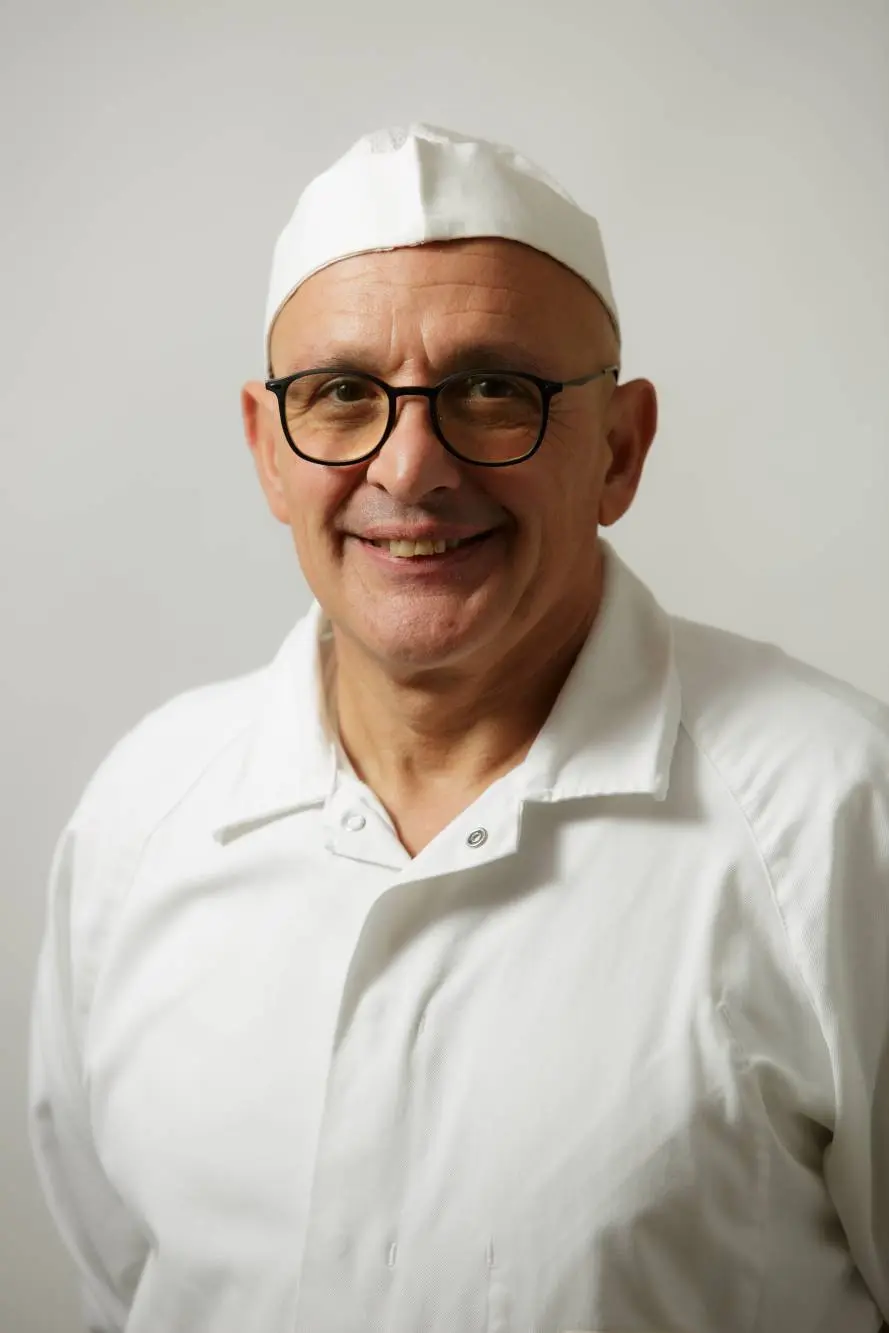 Una persona con gli occhiali, un cappello da cuoco bianco e un impeccabile camice da cuoco, tipico dell'Azienda Carni, sorride alla telecamera su uno sfondo semplice.