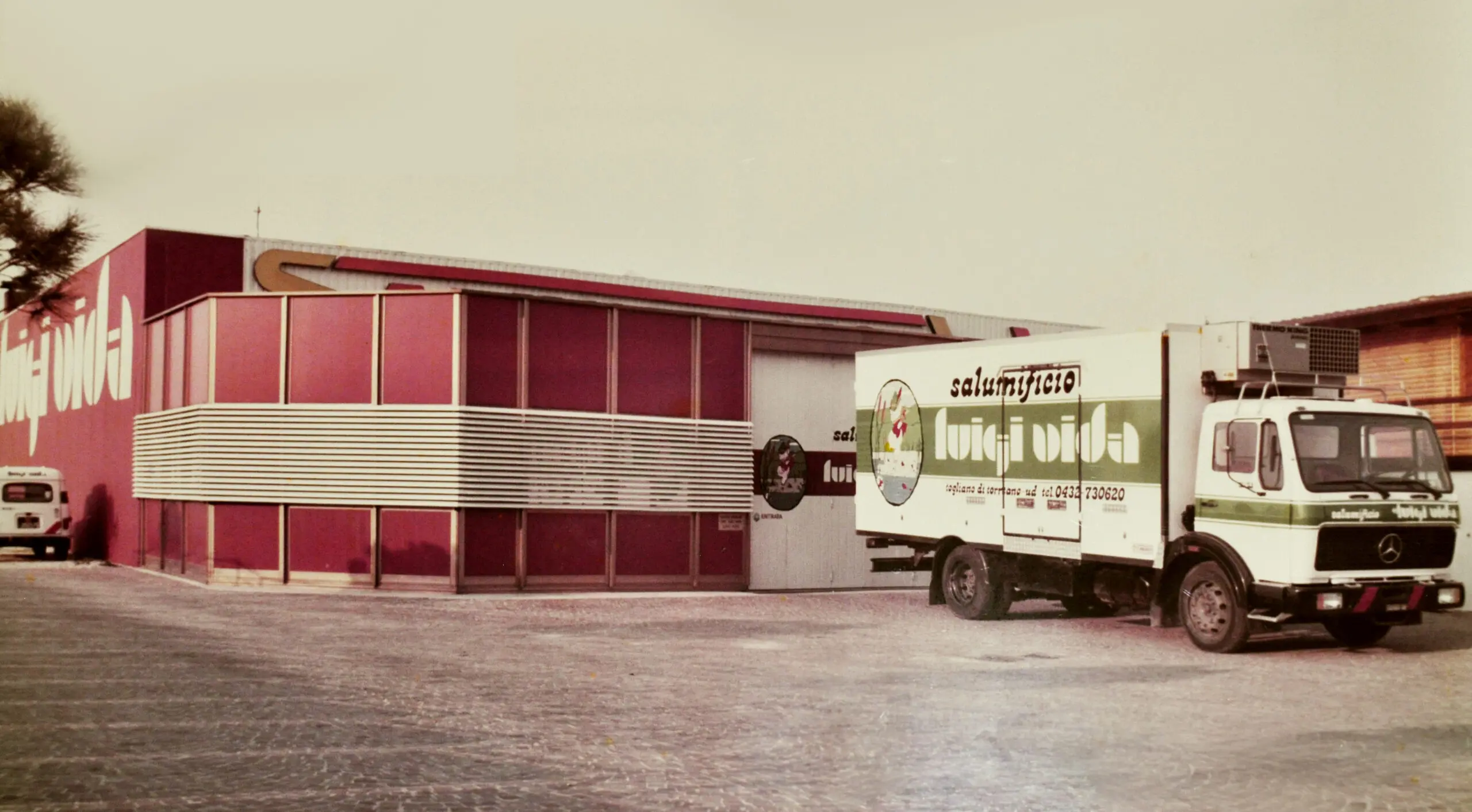 Un camion d'epoca con il marchio "Lacticivora" è parcheggiato fuori da un magazzino rosso e bianco. L'edificio ha un design a strisce e solido sulla parte anteriore. La scena sembra essere in una zona industriale.