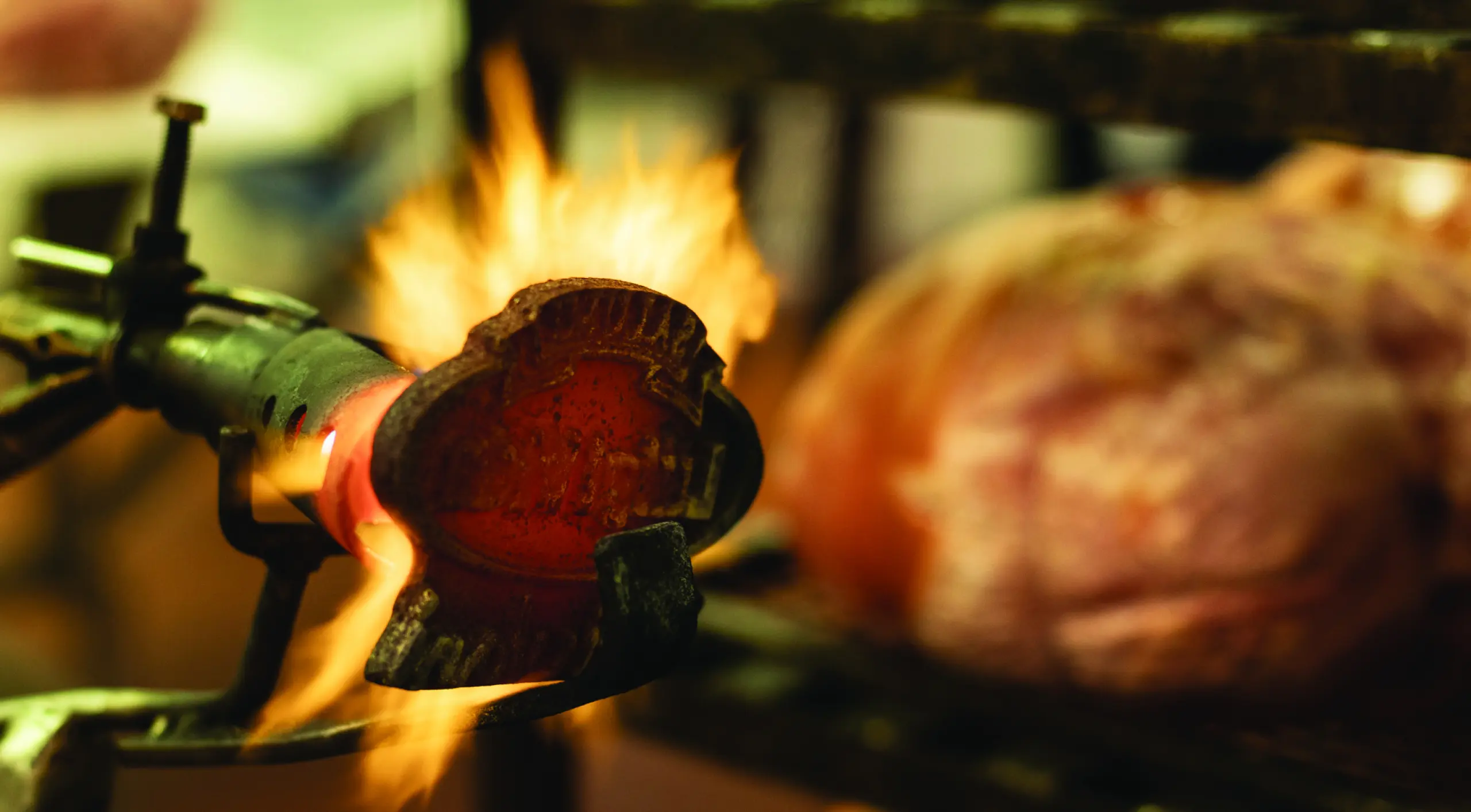 Un primo piano di una torcia con fiamme che riscaldano un ferro da marchiatura. Sullo sfondo, c'è un'immagine sfocata di carne su una griglia. La scena suggerisce la preparazione o la cottura del cibo, con l'attenzione rivolta al bagliore infuocato del ferro da marchiatura.