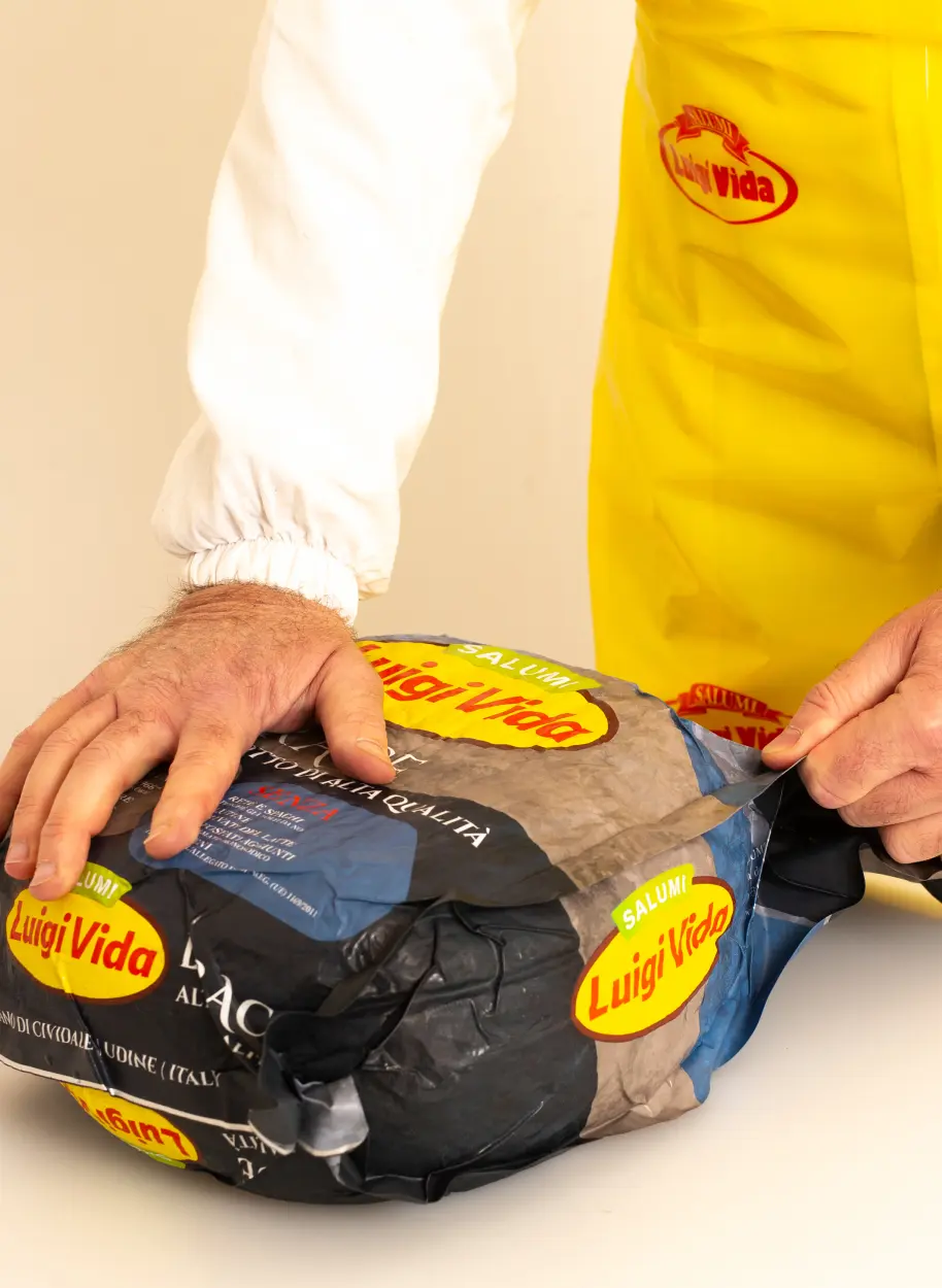 Una persona che indossa un grembiule giallo sta impacchettando un pacco con l'etichetta "Luigi Vida Salumi". Il pacco sembra contenere carne stagionata o un prodotto simile. La persona sta usando entrambe le mani per fissare la confezione.