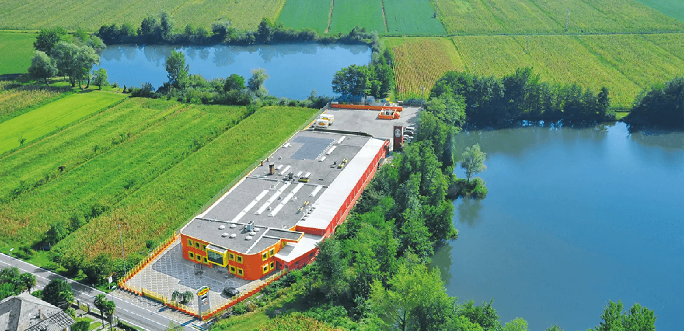 Vista aerea di un grande edificio rettangolare con una facciata colorata, situato tra due piccoli laghi e circondato da rigogliosi campi verdi. Una strada costeggia il bordo della proprietà. Gli alberi costeggiano i corsi d'acqua e i campi.