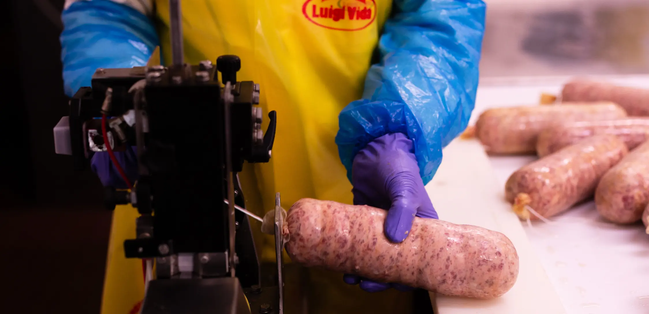 Un lavoratore che indossa guanti blu, un grembiule giallo e maniche blu maneggia una salsiccia in una macchina per la lavorazione della carne. Diverse salsicce sono sul tavolo sullo sfondo.