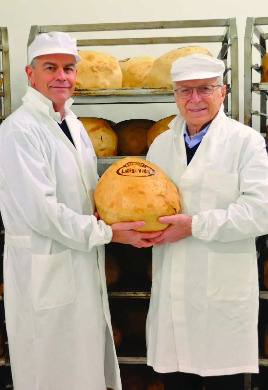 Due uomini in uniforme bianca e berretto sono in piedi davanti a scaffali pieni di grandi forme di formaggio. Sorridono e tengono in mano una forma rotonda di formaggio con inciso "Luigi Guffanti".
