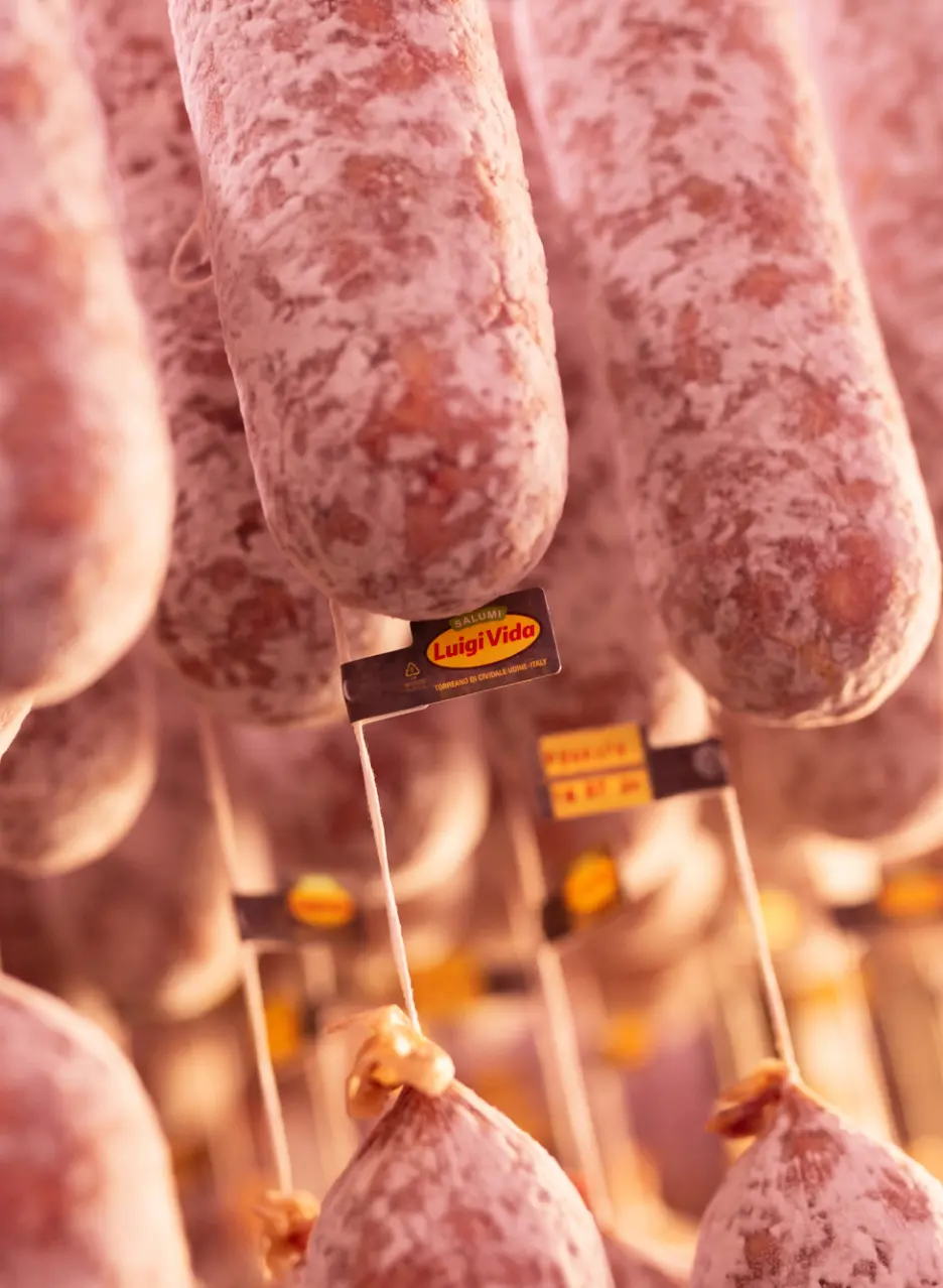 Un primo piano di diverse salsicce appese, con un'etichetta con la scritta "Luigi Vida" attaccata a una. Le salsicce hanno un aspetto rustico e strutturato, che suggerisce che si tratti di salumi. Lo sfondo è leggermente sfocato, esaltando la messa a fuoco in primo piano.
