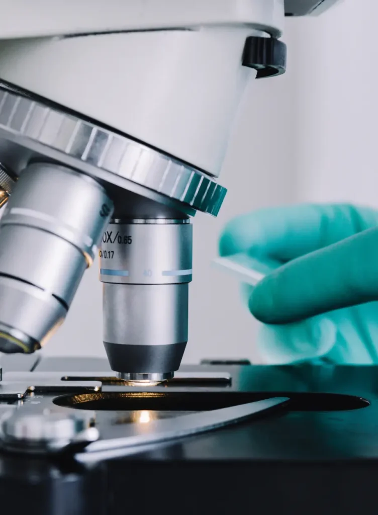 Primo piano di un microscopio con una mano guantata che regola un vetrino sul tavolino. La lente e la luce del microscopio sono focalizzate sul vetrino, indicando un ambiente scientifico o di laboratorio.
