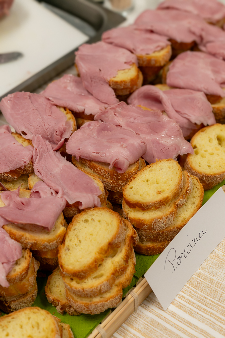 Fette di baguette condite con sottili fette di porchetta rosa e saporita lingua cotta sono disposte in file su un vassoio. Accanto al cibo è posto un piccolo cartellino con la scritta "Porchina".